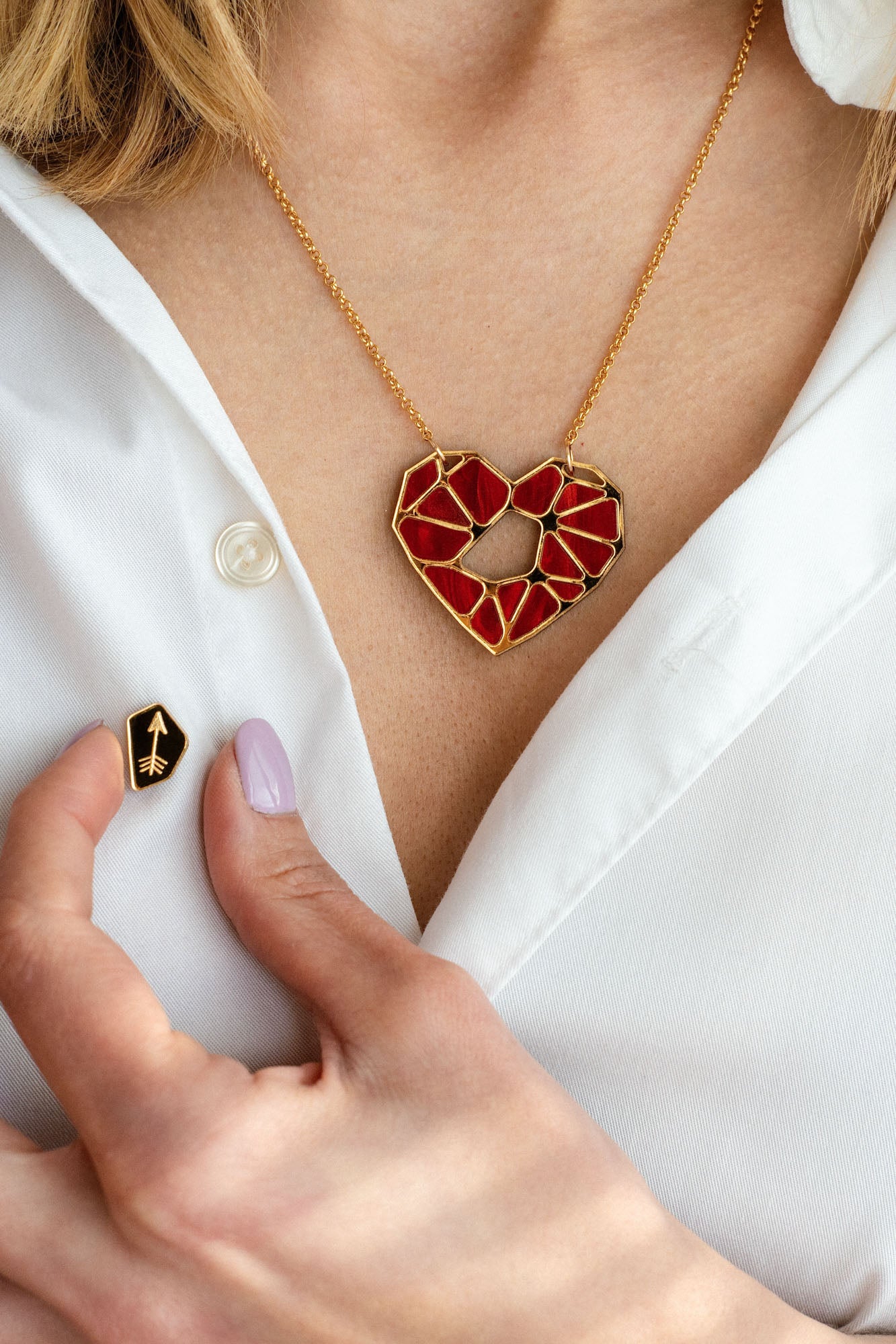 A person wearing a white shirt displays the handcrafted Fragment of Love Necklace featuring a red, geometric heart pendant and holds a small black pin with a gold arrow design. Only the lower face and hand are visible.