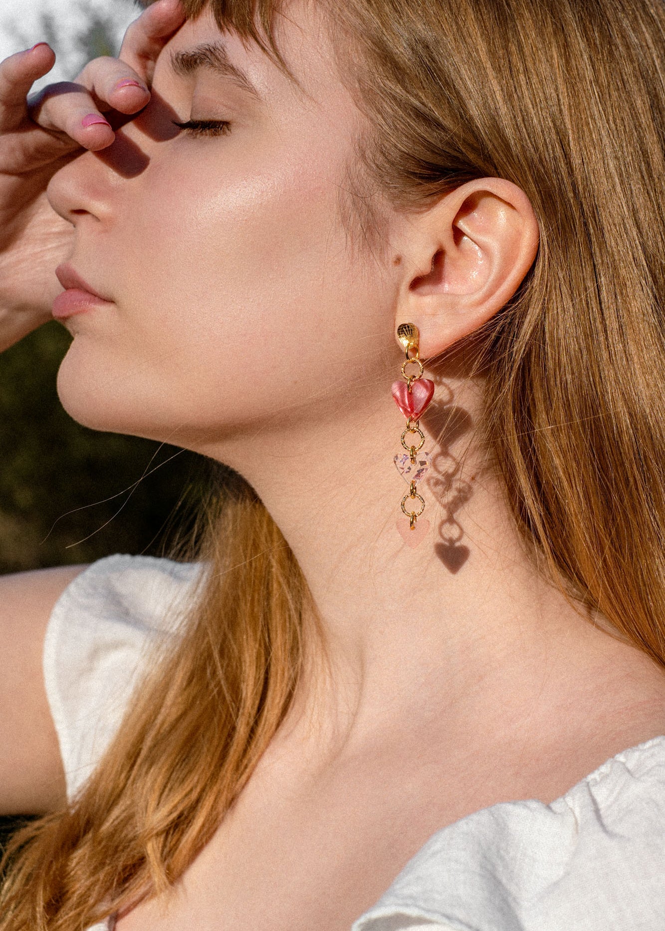 A woman with long brown dreadlocks and gold Heart Chain Earrings gazes to the left. She wears a cream-colored blouse and stands against a backdrop of pink curtains, highlighting soft pink tones throughout the scene.