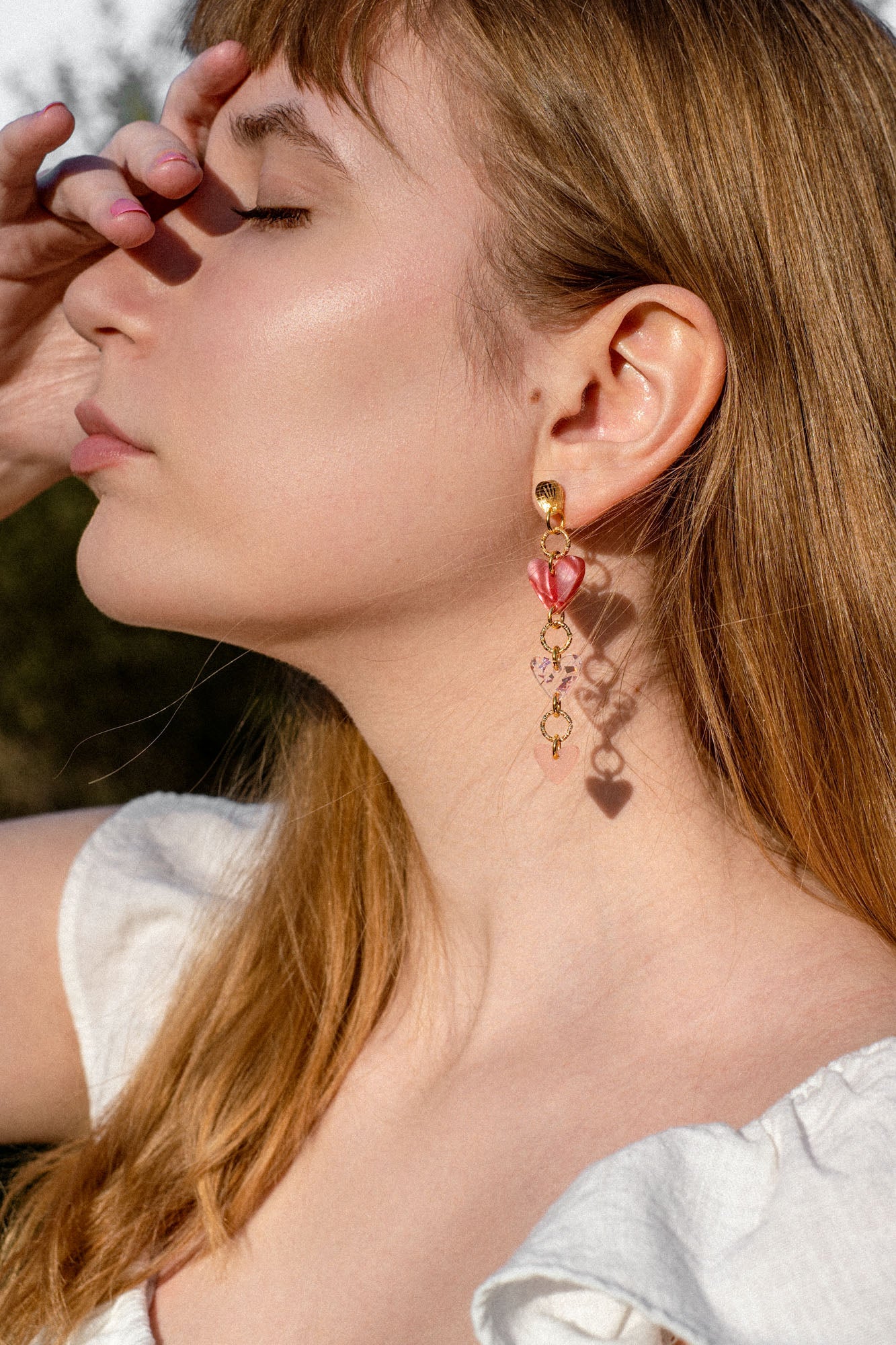 A woman with long brown dreadlocks and gold Heart Chain Earrings gazes to the left. She wears a cream-colored blouse and stands against a backdrop of pink curtains, highlighting soft pink tones throughout the scene.