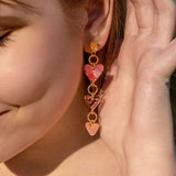 Close-up of a person wearing Heart Chain Earrings in gold and pink tones. They have braided hair and a light-colored top, with an out-of-focus background. Designed for allergy-free comfort, these earrings add a playful touch to any look.
