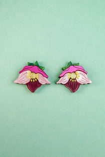 Two colorful Hellebore Stud Earrings, shaped like stylized pink flowers with green leaves and yellow centers, are displayed on a light turquoise background—perfect handcrafted floral jewelry for unique accessories lovers.