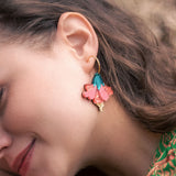 A woman with wavy brown hair wears vibrant Hibiscus Earrings—gold hoops adorned with pink, teal, and gold floral charms. She is smiling softly, her tropical statement green patterned clothing visible.