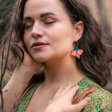 A woman with wavy brown hair and closed eyes stands outdoors, wearing a green patterned dress and vibrant Hibiscus Earrings, touching her face and neck gently, with a tattoo visible on her forearm.