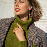 Woman wearing a green turtleneck sweater and plaid coat, accessorized with handcrafted Hummingbird Earrings and a brooch, poses outdoors against a light textured wall, touching her collar and looking off to the side.