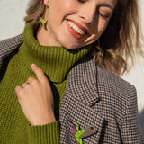 A smiling woman in a green turtleneck and plaid blazer stands in sunlight, eyes closed and head tilted. She wears bird-shaped earrings and a handcrafted Hummingbird Brooch, all made from premium materials.