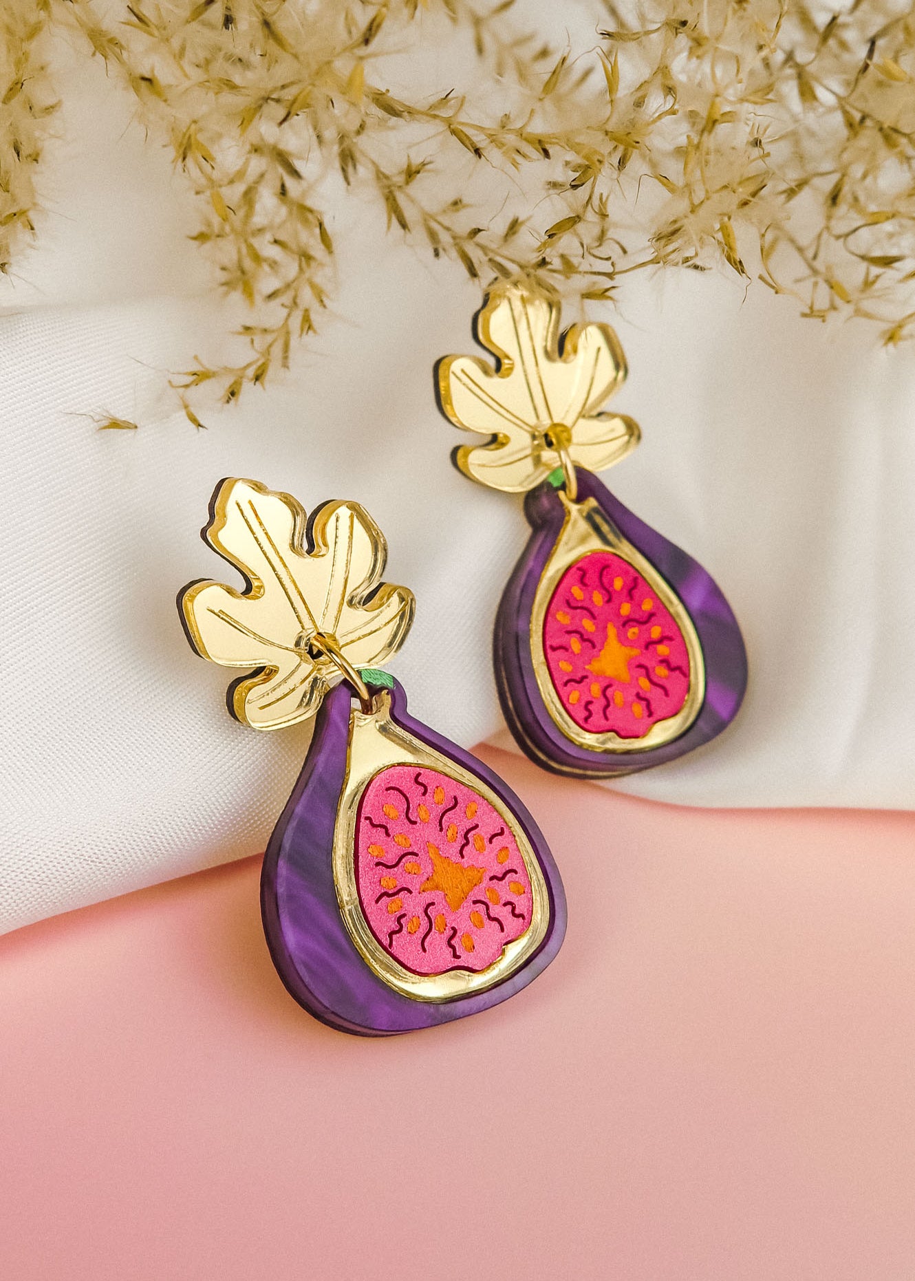 A pair of handcrafted Fig Earrings with gold mirror leaves, purple edges, and a pink center, displayed on a soft white and pink background with dried flowers.
