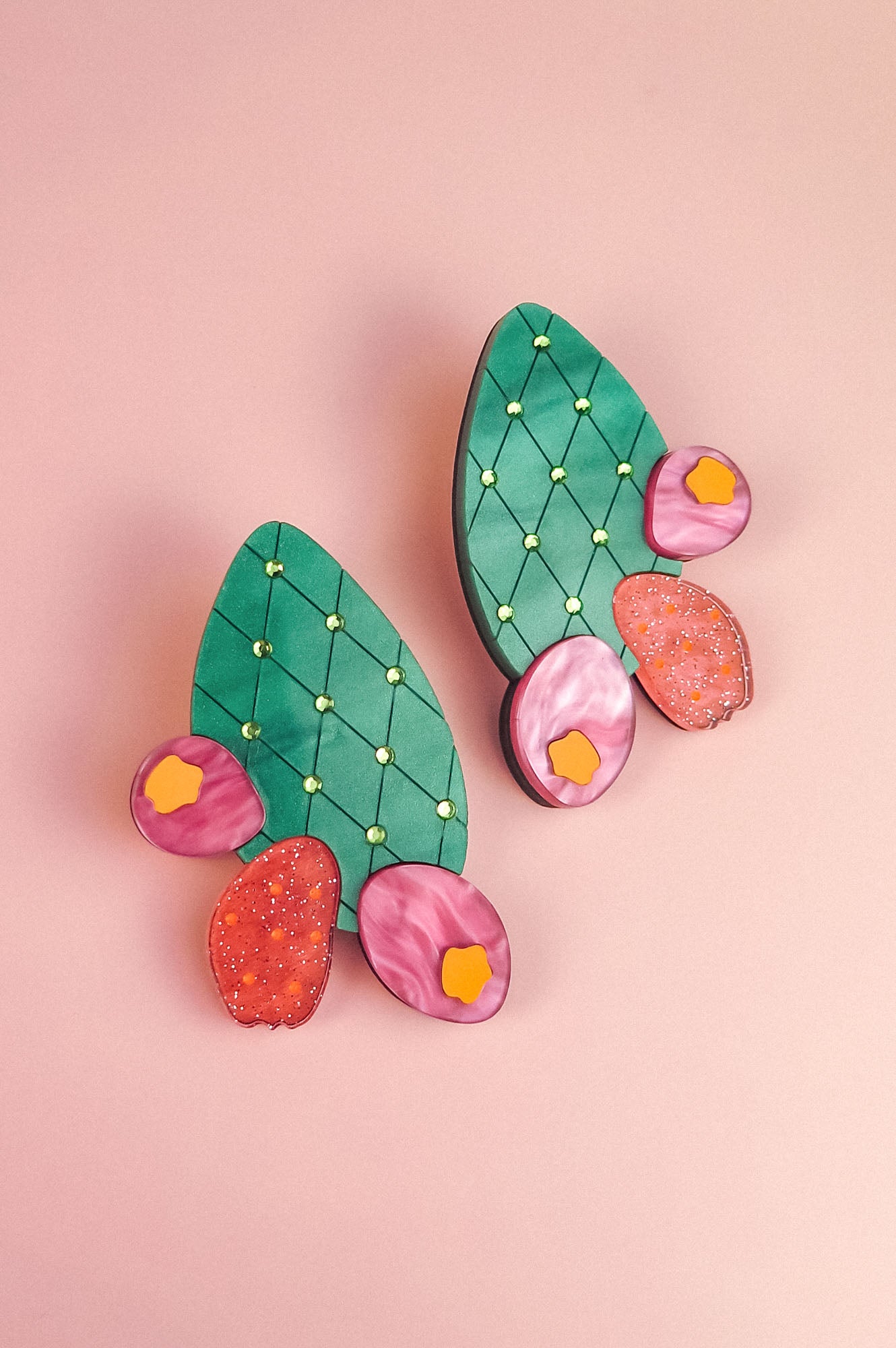 A pair of statement earrings shaped like green opuncia cactus pads with pink and orange flowers, decorated with small yellow dots, displayed on a light pink background.