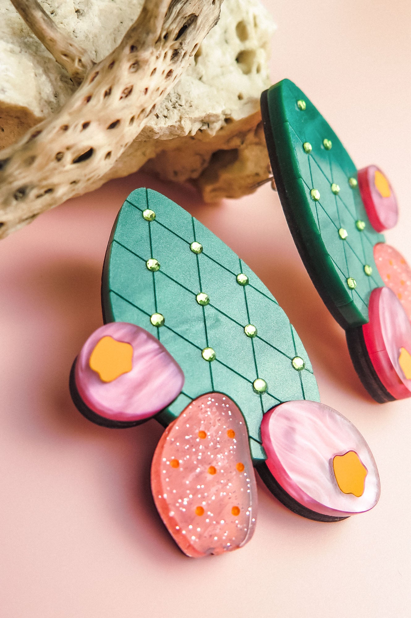 A close-up of colorful, handmade statement earrings shaped like opuncia cactus pads with green, pink, and orange sections, lying on a pink surface next to a textured piece of driftwood.