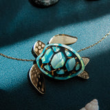 A gold and turquoise Sea Turtle Necklace pendant on a chain sits on a teal surface, surrounded by seashells and lit by soft sunlight—a stunning piece of handcrafted jewelry perfect for ocean lovers.