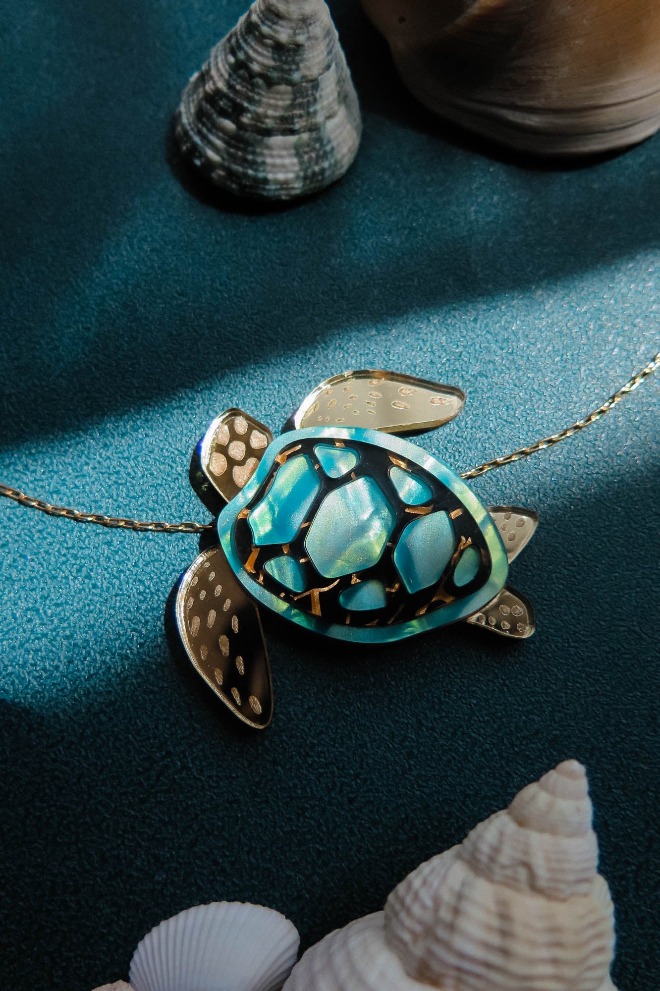 A gold and turquoise Sea Turtle Necklace pendant on a chain sits on a teal surface, surrounded by seashells and lit by soft sunlight—a stunning piece of handcrafted jewelry perfect for ocean lovers.