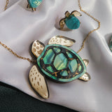 A handcrafted gold jewelry set shaped like sea turtles with green and cream details is displayed on white satin fabric. The set includes a Sea Turtle Necklace, a ring, and earrings. A shell is placed nearby.