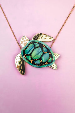 This handcrafted jewelry piece is a gold chain Sea Turtle Necklace, featuring a turtle pendant with a green and black patterned shell and elegant gold accents, beautifully set against a pink background.