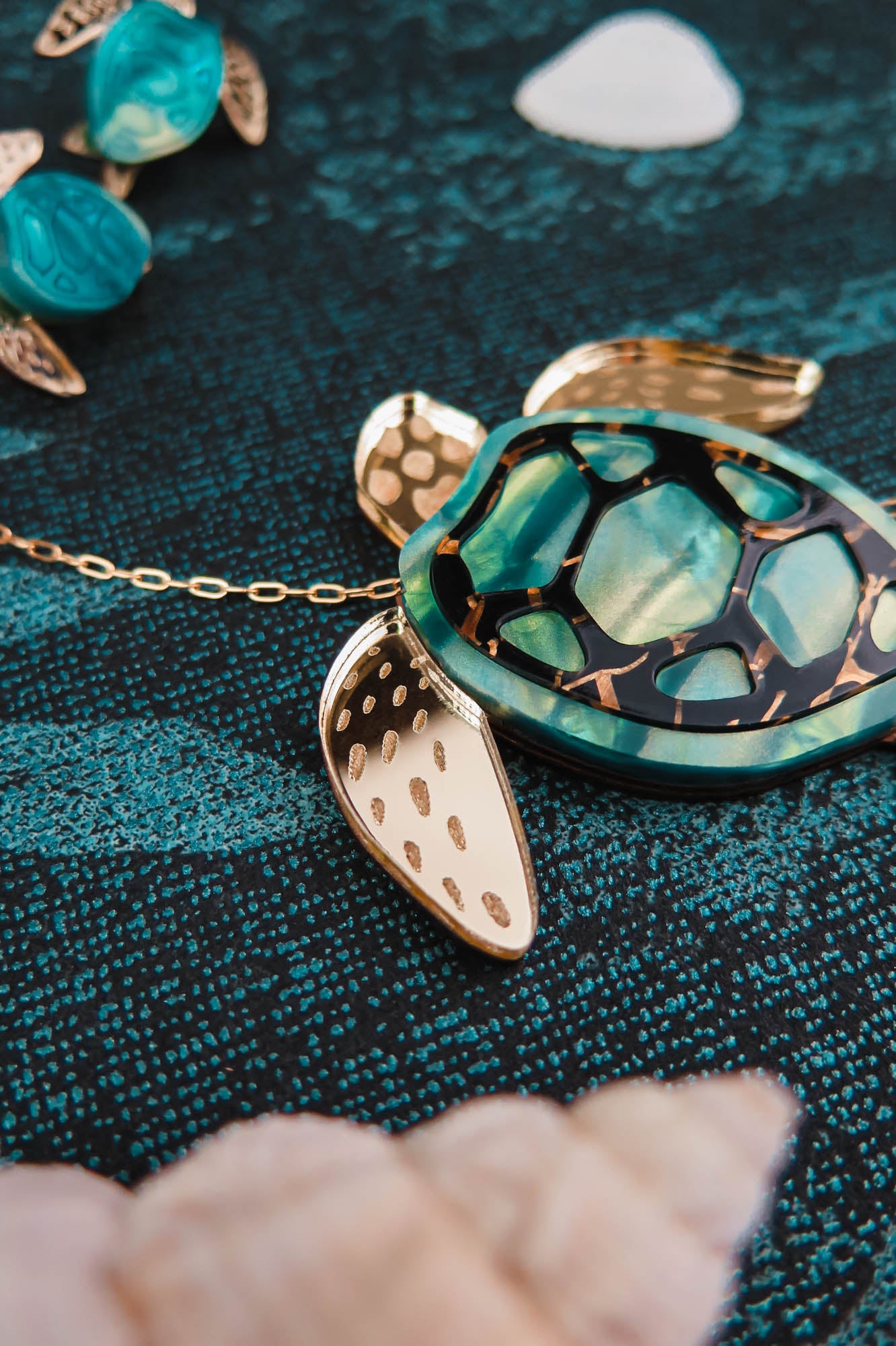 A close-up of a gold and turquoise Sea Turtle Necklace lies on a textured blue surface, with seashells and other handcrafted jewelry featuring turtle-themed designs nearby.