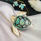 A gold and green Sea Turtle Necklace pendant, handcrafted jewelry with intricate details, is displayed on silky white fabric alongside a matching smaller turtle necklace in the background and a seashell nearby.