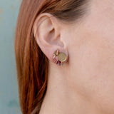 Close-up of a person with light skin and reddish-brown hair wearing a small, round, gold Minimalist Geometric Stud Earring featuring a wavy, pink and green patterned design on their left ear against a light blue background.