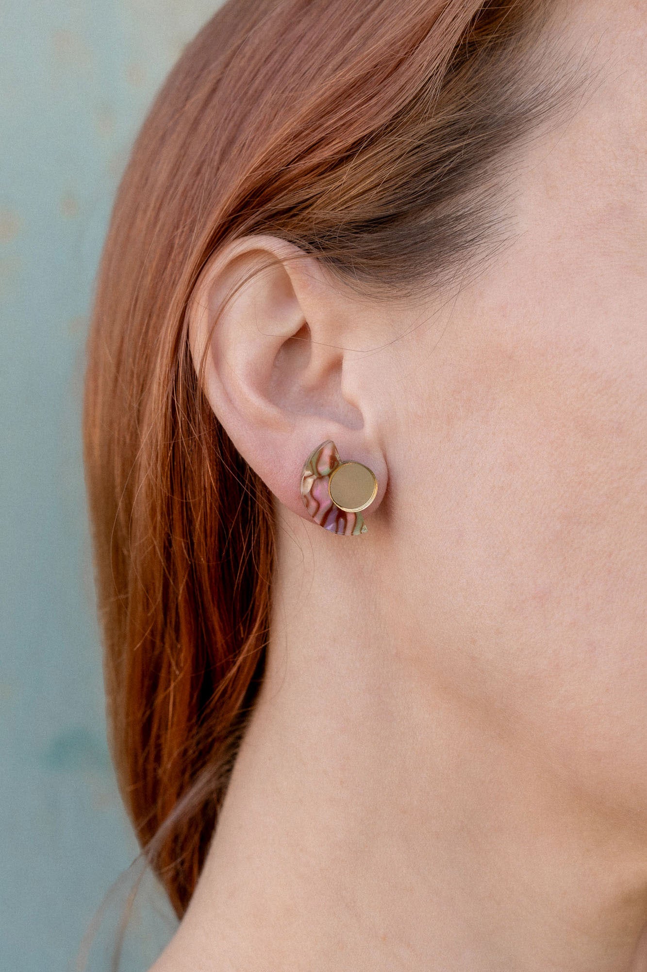 Close-up of a person with light skin and reddish-brown hair wearing a small, round, gold Minimalist Geometric Stud Earring featuring a wavy, pink and green patterned design on their left ear against a light blue background.