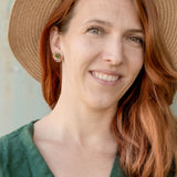 A woman with long auburn hair wears minimalist geometric stud earrings, a wide-brimmed straw hat, and a green dress, smiling softly while standing in front of a neutral-colored wall.
