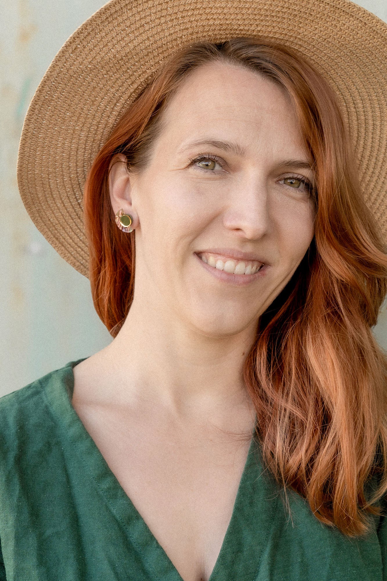 A woman with long auburn hair wears minimalist geometric stud earrings, a wide-brimmed straw hat, and a green dress, smiling softly while standing in front of a neutral-colored wall.