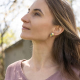A woman with long brown hair wearing a mauve blouse and minimalist jewelry—heart-shaped earrings—looks upward while gently touching her hair; soft natural light and a blurred outdoor background.