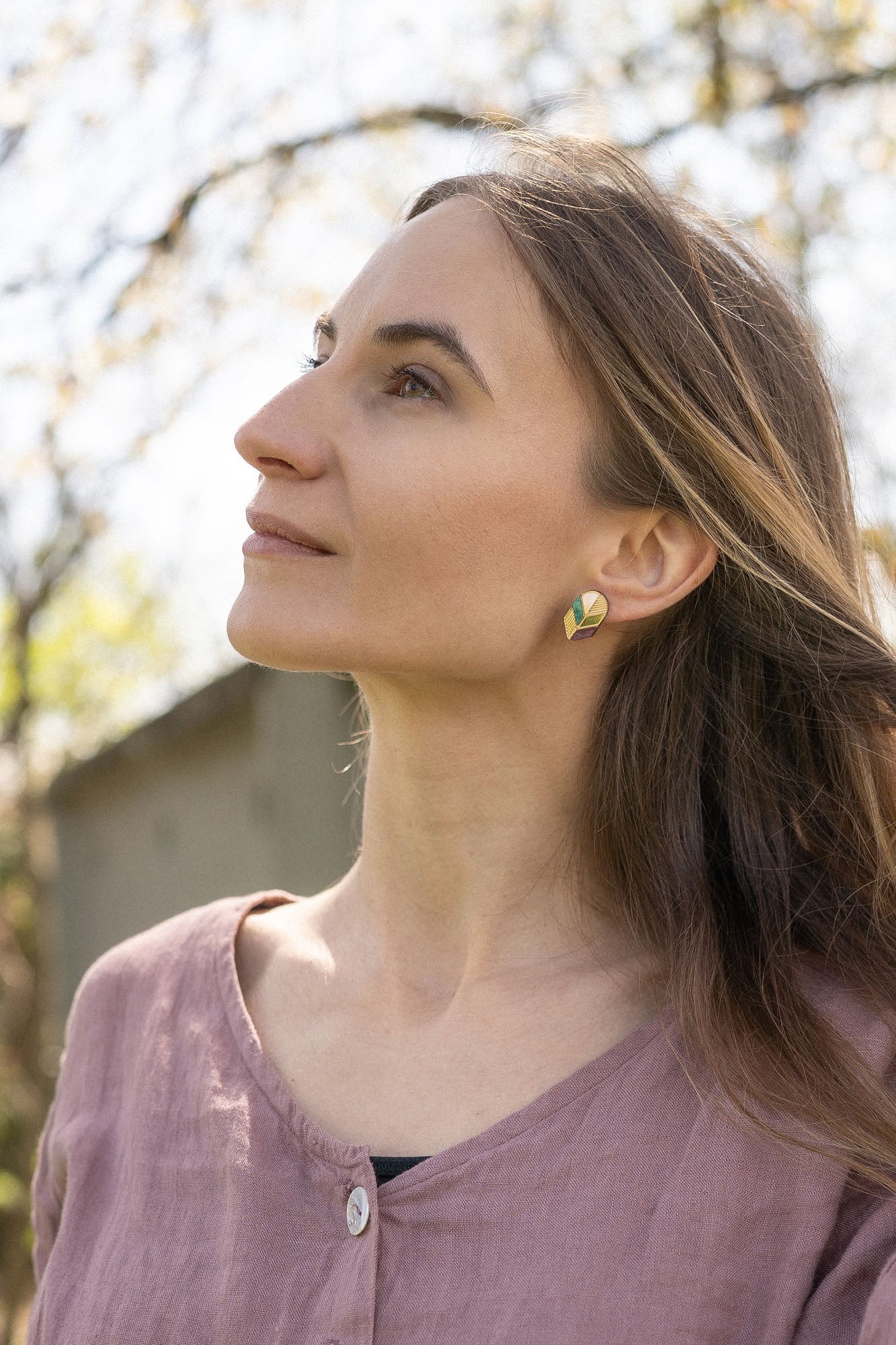 A woman with long brown hair wearing a mauve blouse and minimalist jewelry—heart-shaped earrings—looks upward while gently touching her hair; soft natural light and a blurred outdoor background.
