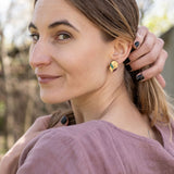 A woman with light brown hair in a low ponytail stands outdoors, wearing a mauve blouse and minimalist jewelry—gold leaf studs that catch the light. She looks over her shoulder at the camera, smiling softly, with one hand touching her hair.