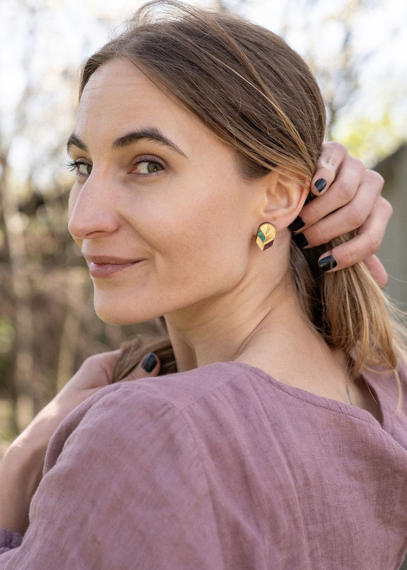 A woman with light brown hair in a low ponytail stands outdoors, wearing a mauve blouse and minimalist jewelry—gold leaf studs that catch the light. She looks over her shoulder at the camera, smiling softly, with one hand touching her hair.