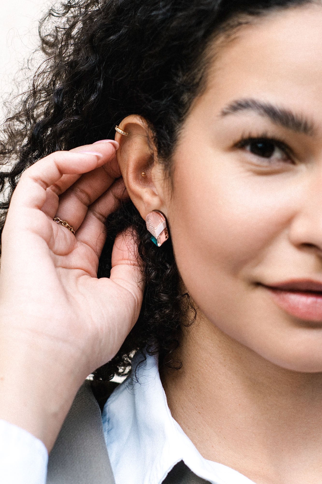 A close-up of a woman with curly hair touching her ear, showcasing minimalist jewelry with geometric earrings and a gold ear cuff. She is wearing a white collared shirt and light-colored jacket, smiling slightly.