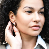 A woman with curly dark hair gazes to the side, gently touching her ear to show off handmade minimalist jewelry—delicate leaf studs. She wears a white collared shirt and a light gray jacket.