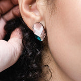 Close-up of a person’s ear wearing a geometric, handmade earring with metallic, pink, and teal segments. The look is completed with minimalist jewelry: a small gold stud, gold ear cuff, and curly hair visible near the ear.