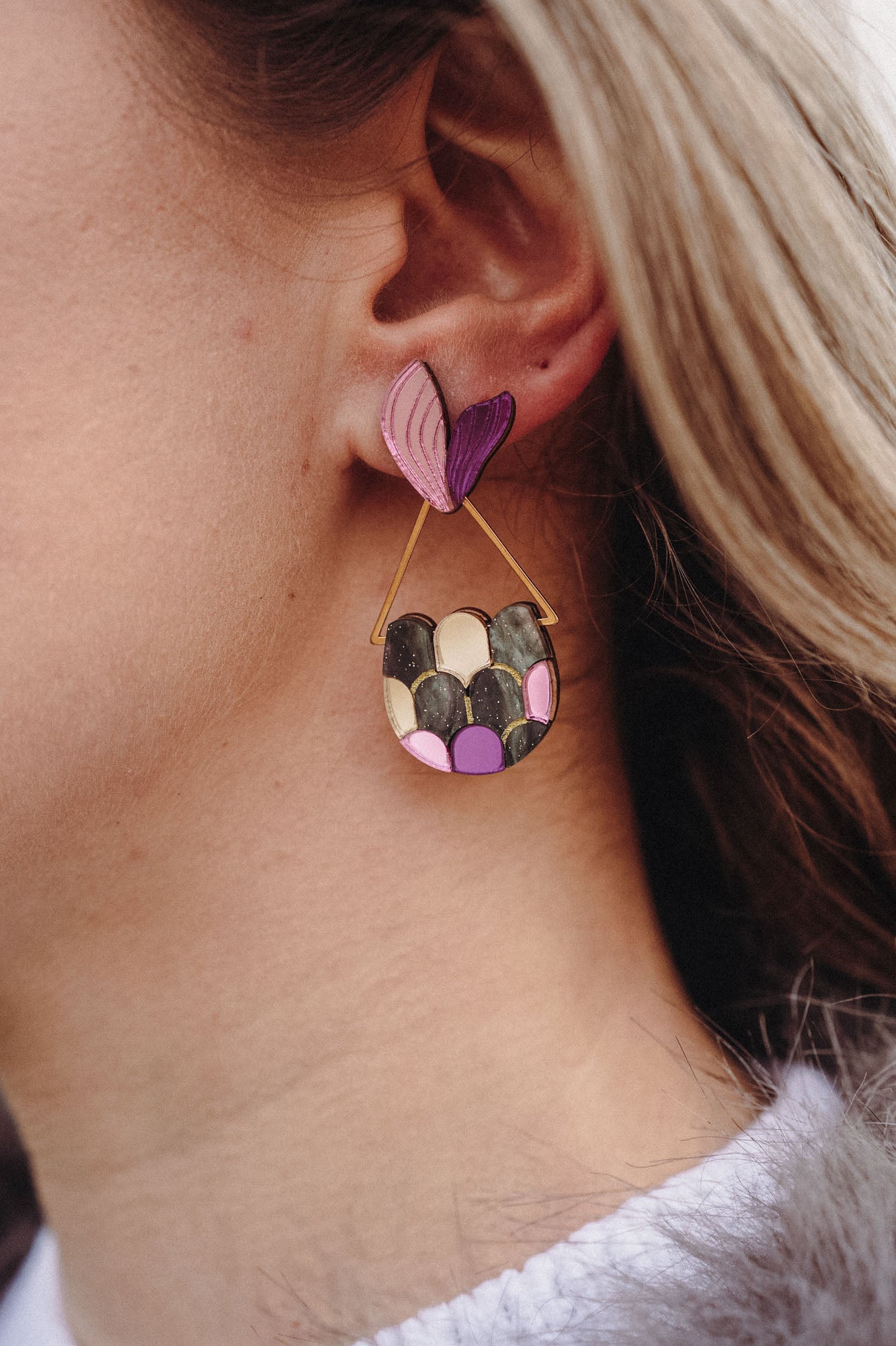 Close-up of a woman's ear wearing playful Mermaid Tail Earrings with a pink and purple heart-shaped stud and a semi-circular dangle in green, pink, and purple—a perfect accessory for mermaid lovers. The woman has blonde hair and wears a white top.