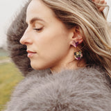 A woman with wavy blonde hair, wearing a fluffy gray coat and playful Mermaid Tail Earrings, stands outdoors with her eyes closed and one hand in her hair. The background is grassy with blurred trees—perfect for mermaid lovers.