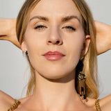 A woman with long blonde hair and a patterned dress faces the camera with her hands behind her head, wearing handcrafted earrings in bright sunlight.