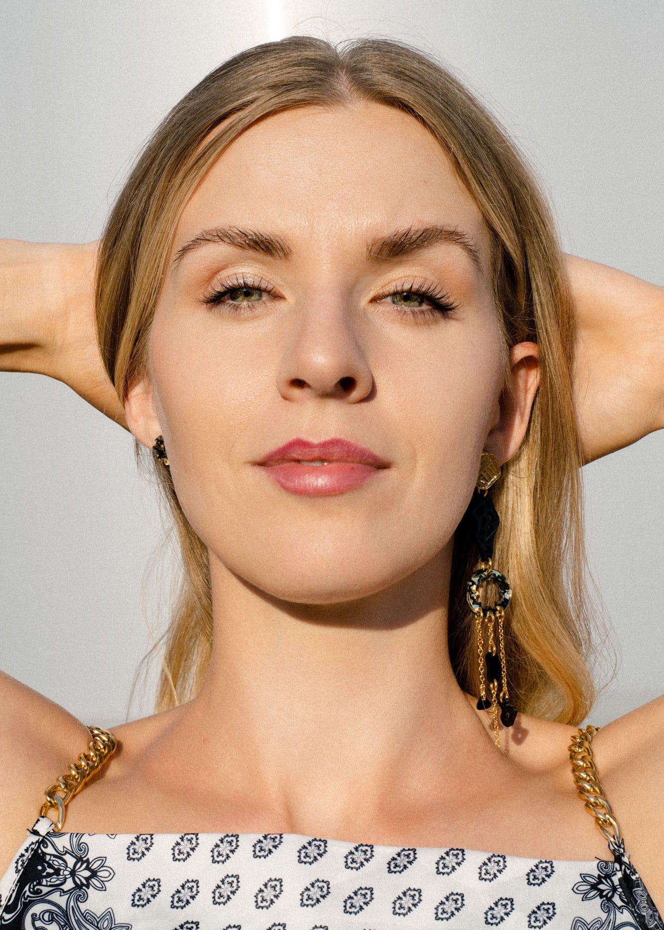 A woman with long blonde hair and a patterned dress faces the camera with her hands behind her head, wearing handcrafted earrings in bright sunlight.