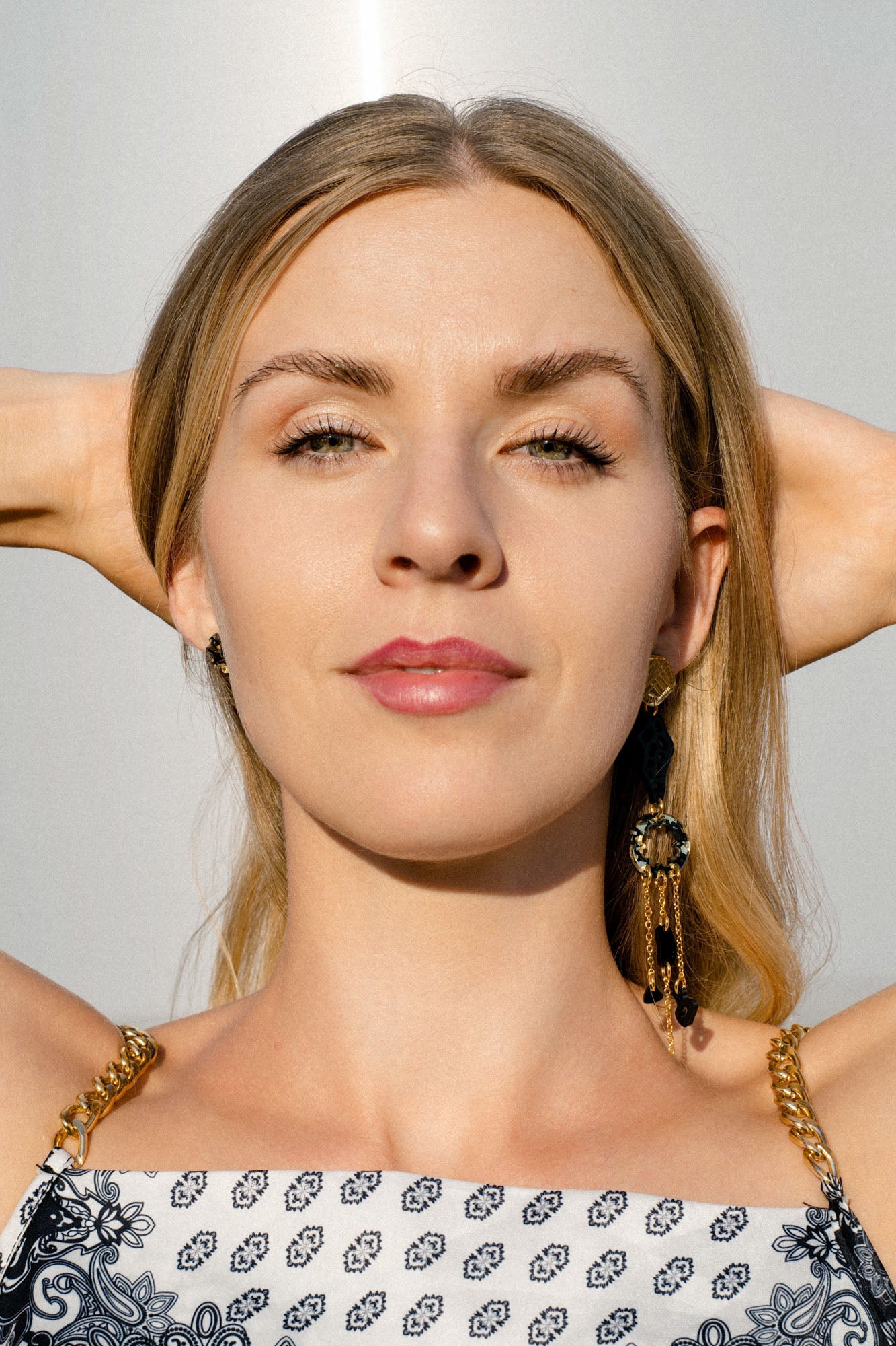 A woman with long blonde hair and a patterned dress faces the camera with her hands behind her head, wearing handcrafted earrings in bright sunlight.