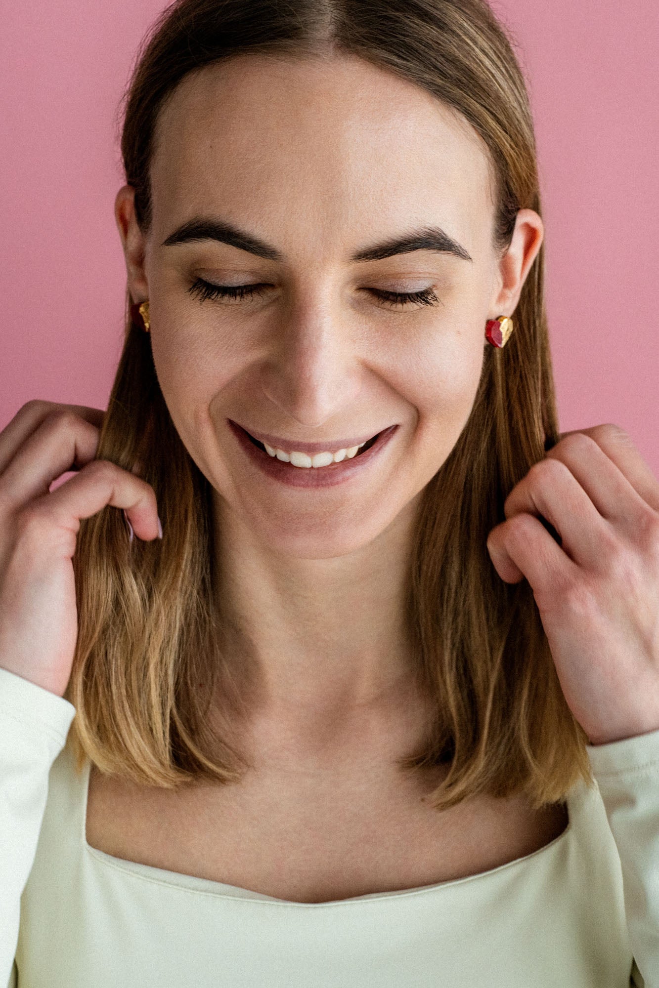 A woman with shoulder-length brown hair and a white top smiles with her eyes closed against a pink background, touching her hair with both hands, while wearing delicate handcrafted earrings.