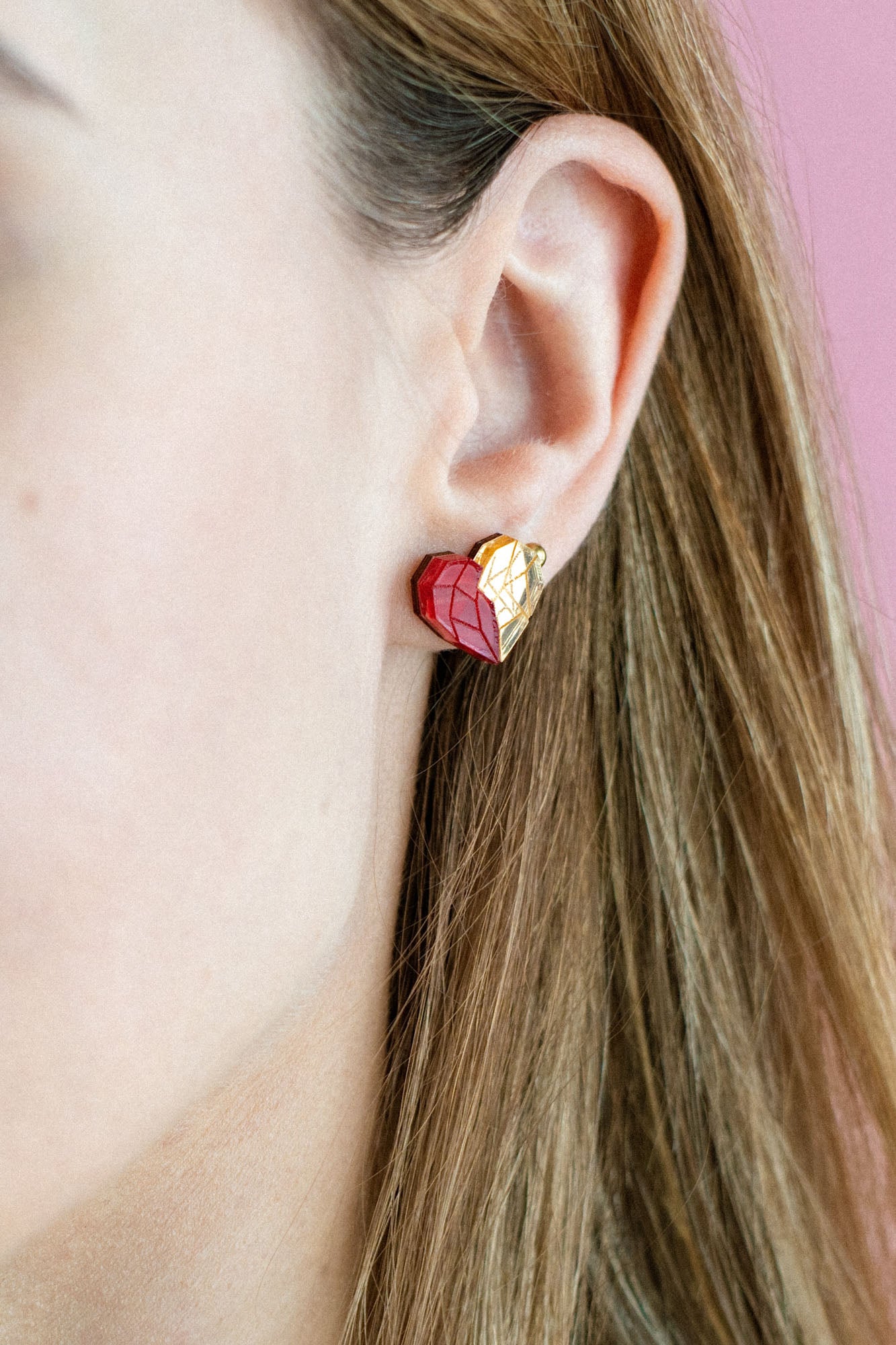 A close-up of a woman with light brown hair in a cream top, showing off romantic jewelry—a red and gold heart-shaped handcrafted earring. The pink background highlights the elegant design, with only the right side of her face visible.