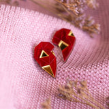 A pair of handcrafted geometric red and gold earrings shaped like hearts rest on textured pink fabric, with small dried flowers placed nearby—a perfect romantic gift.