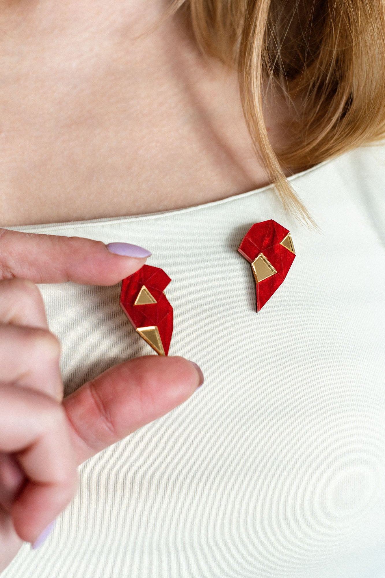 A person with long hair wears a cream top, holding a red and gold geometric One Love Halves Pin shaped like half a broken heart—the other handcrafted half is pinned to their shirt near the collarbone. Perfect as a romantic gift.
