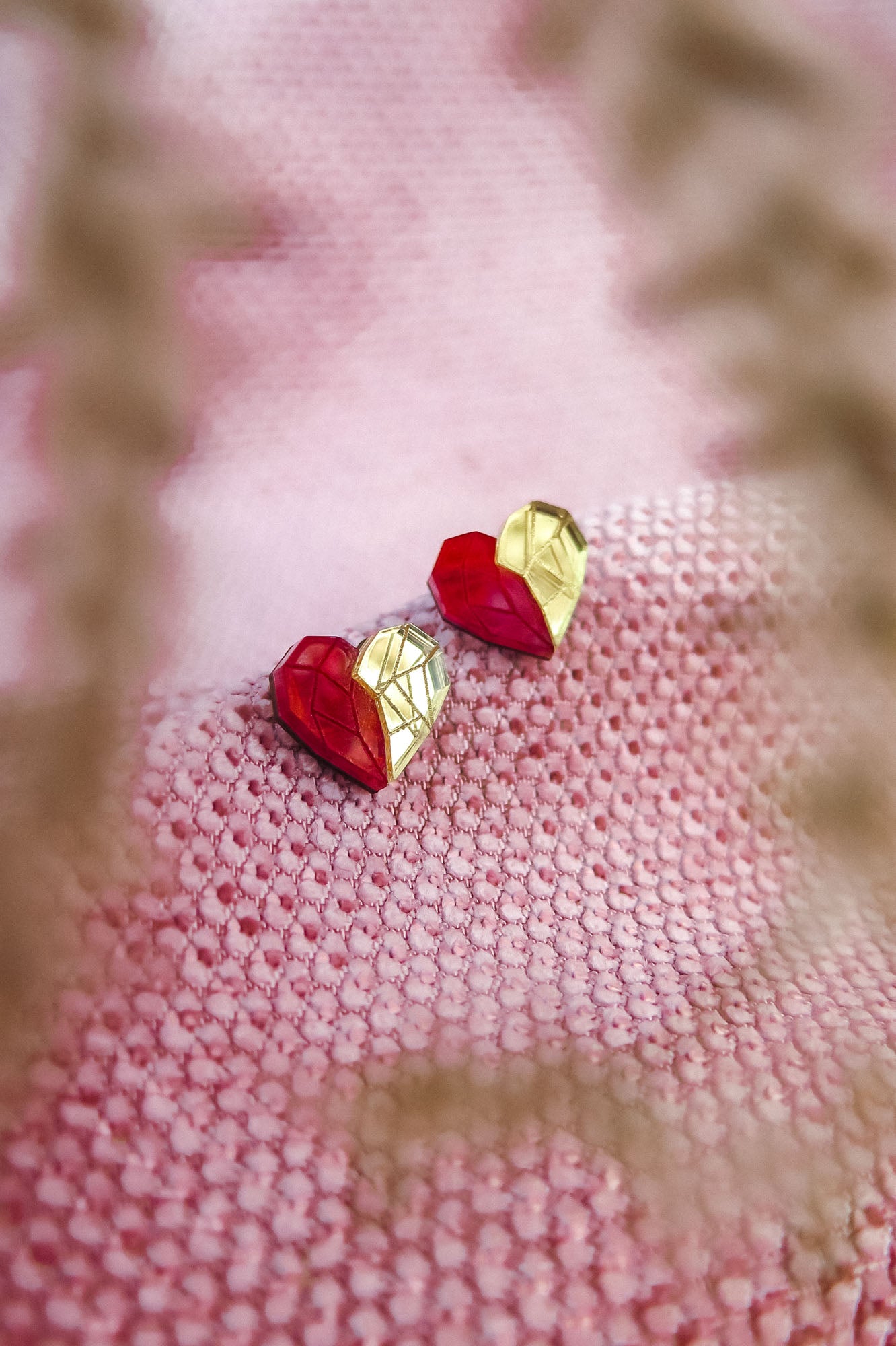 A pair of handcrafted red and gold geometric heart-shaped earrings rest on a textured pink fabric, with blurry plant stems in the foreground—perfect romantic jewelry for any occasion.
