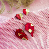 Red and gold geometric heart studs and pins, perfect as romantic jewelry, are displayed on textured pink fabric, with decorative plant stems in the background.