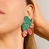 Close-up of a woman's face wearing large, statement earrings shaped like an opuncia cactus with pink and yellow decorative accents. Her hand touches her hair, and her eyes are closed, highlighting the bold design of the accessory.