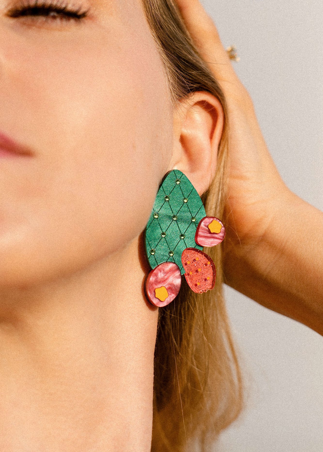Close-up of a woman's face wearing large, statement earrings shaped like an opuncia cactus with pink and yellow decorative accents. Her hand touches her hair, and her eyes are closed, highlighting the bold design of the accessory.