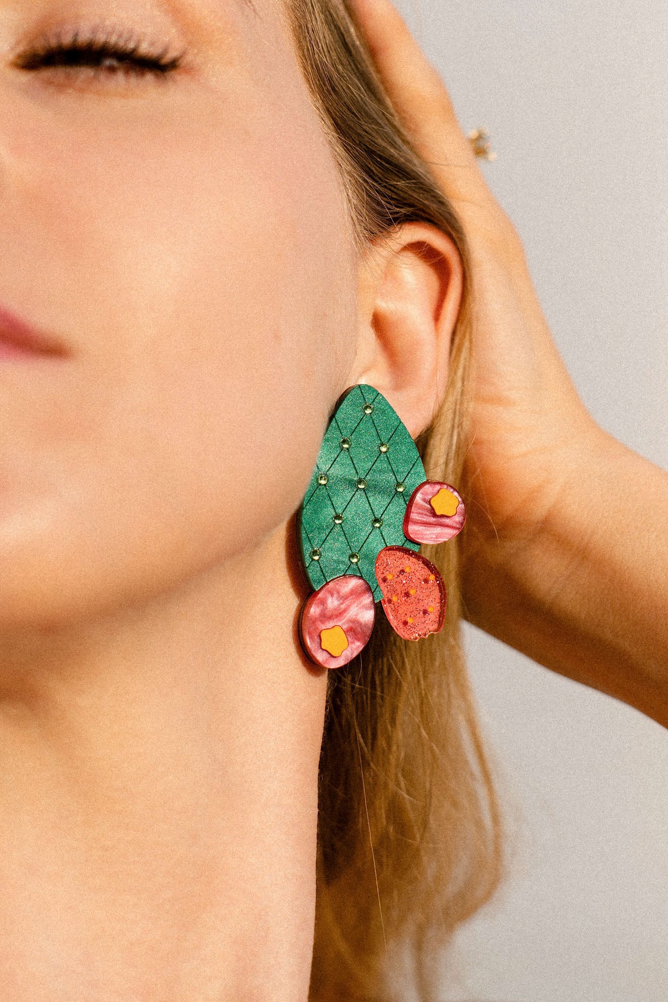 Close-up of a woman's face wearing large, statement earrings shaped like an opuncia cactus with pink and yellow decorative accents. Her hand touches her hair, and her eyes are closed, highlighting the bold design of the accessory.