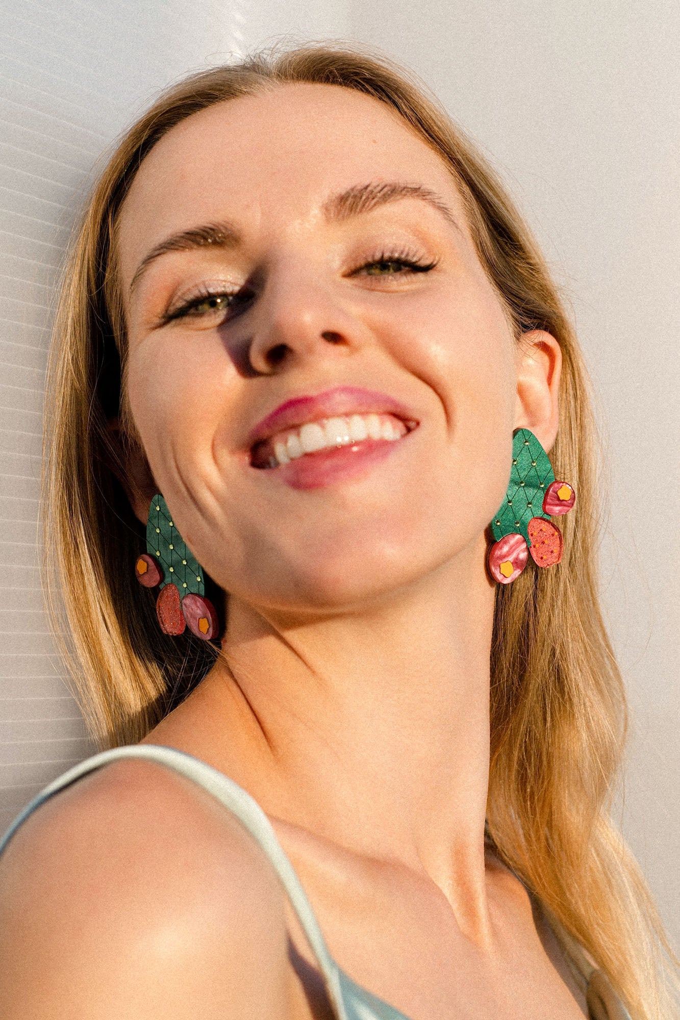 A woman with long blonde hair smiles brightly at the camera, wearing bold green and pink statement earrings featuring an opuncia cactus design, and a light blue top. Sunlight highlights her face and her bold earrings.