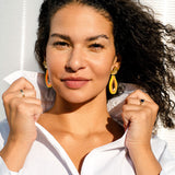 Woman with curly hair and colorful neon acrylic earrings poses confidently, holding the collar of her white shirt. Sunlight shines on her face, highlighting her features. She wears a ring on each hand and stands against a light, textured background.