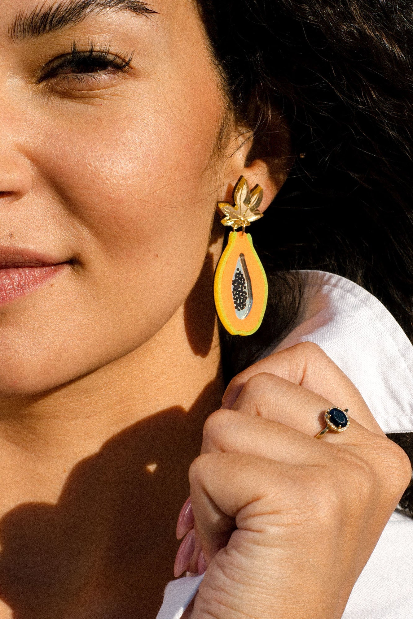 A woman wearing a white shirt shows off a gold ring with a dark blue stone and a colorful Papaya Earring, handcrafted in the shape of a papaya half.