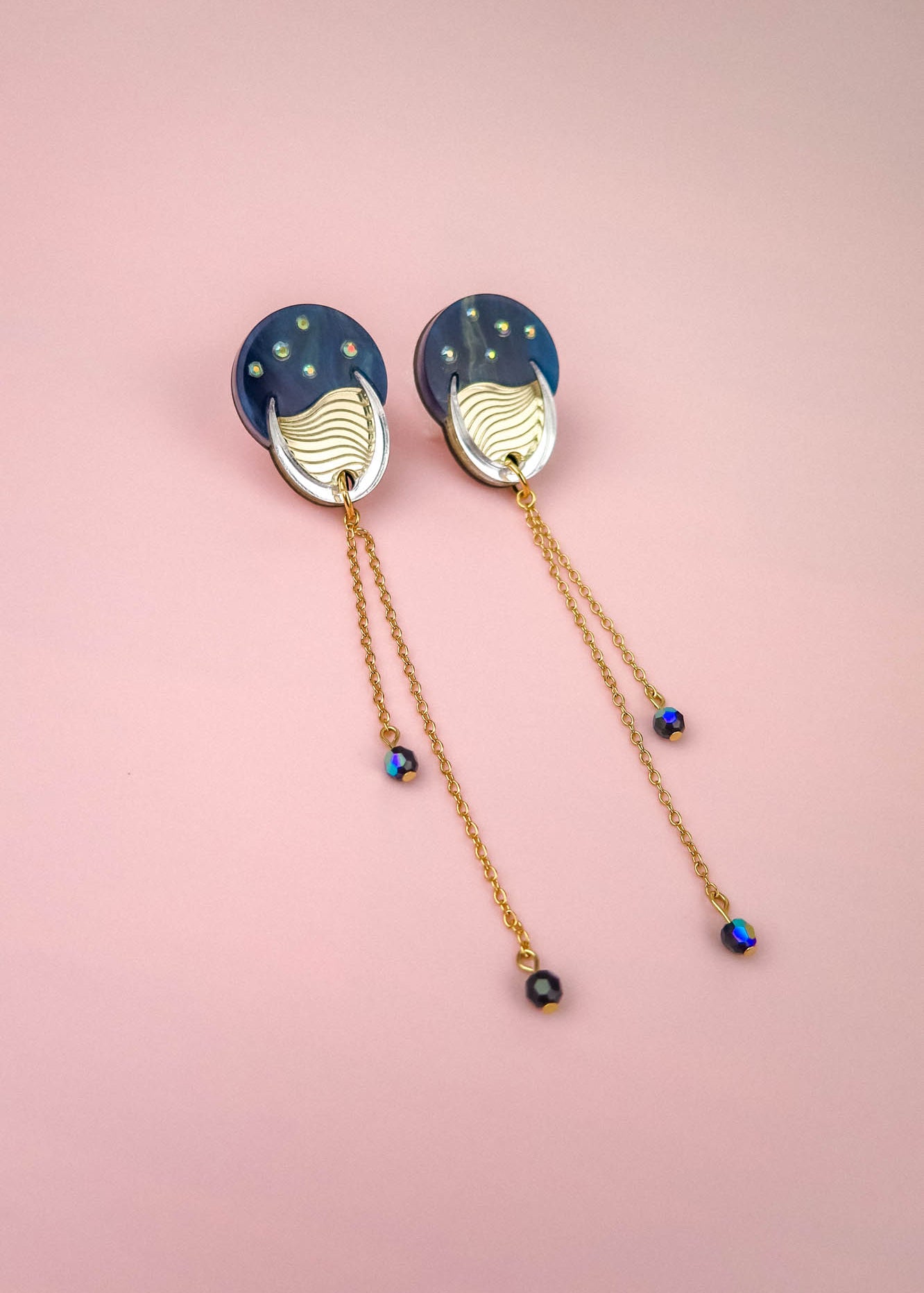 A pair of earrings with blue circular tops reminiscent of the night sky, gold accents, and long gold chains with small blue beads, displayed on a pink background.