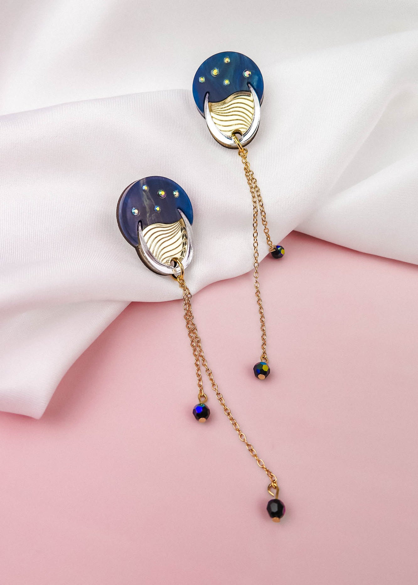 Two gold and blue earrings with dangling chains and beads rest on a white fabric, set against a pink background, evoking a hint of festival magic.