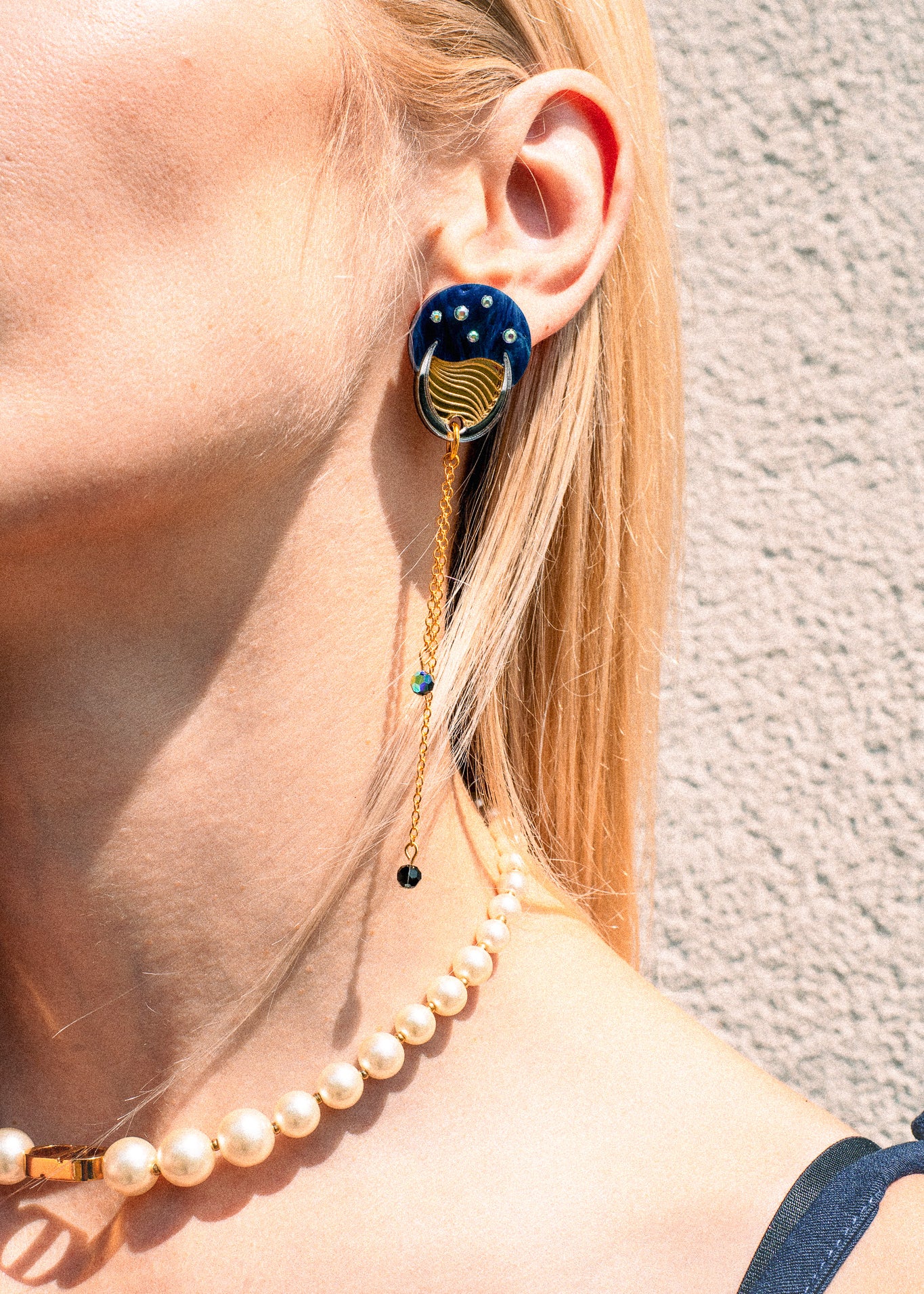 A close-up of a person wearing a blue and gold statement earring with dangling chains that evoke the shimmer of the night sky, paired with a pearl necklace. The person has blonde hair and light skin, and stands against a textured beige wall.