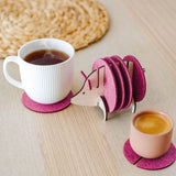 A white cup and a pink cup sit on purple felt coasters next to a wooden hedgehog-shaped coaster holder, with more felt coasters inside it, on a light wooden table—a lovely housewarming gift. A woven mat is in the background.